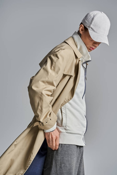 A stylish young man with tattoos wears a trench coat and hat, exuding an air of mystery in a studio setting against a grey background. - Photo, Image