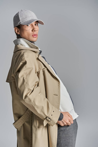 Een stijlvolle man met een opvallende hoed en elegante trench coat poseert zelfverzekerd in een studio tegen een grijze achtergrond. - Foto, afbeelding