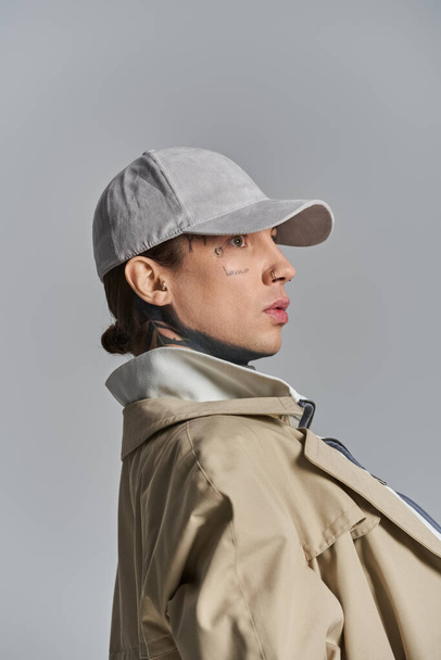 Un jeune homme tatoué pose en toute confiance dans un trench-coat, un chapeau et une veste sur fond de studio gris. - Photo, image