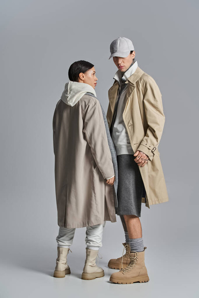A young stylish couple in trench coats standing next to each other in a studio against a grey background. - Photo, Image