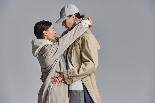 Молода, стильна пара стоїть пліч-о-пліч в траншеї пальто на сірому студійному фоні. - Фото, зображення