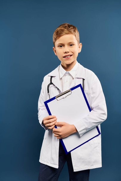 Ένα έφηβο αγόρι σε ένα παλτό γιατρού κρατώντας ένα πρόχειρο σε ένα μπλε φόντο. - Φωτογραφία, εικόνα