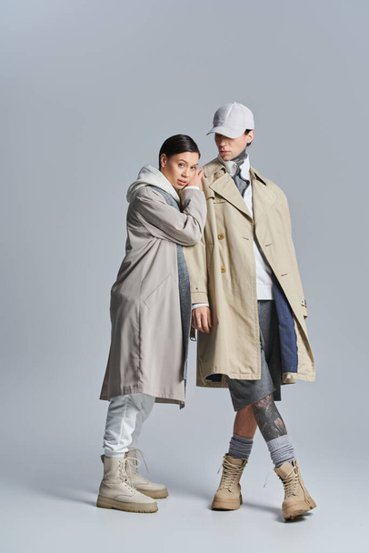 Una pareja joven y elegante se encuentra lado a lado en abrigos de trinchera, exudando sofisticación y elegancia en un estudio sobre un fondo gris. - Foto, imagen