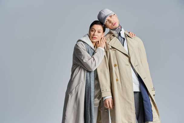 Una pareja joven y elegante se encuentra lado a lado, exudando elegancia en abrigos de trinchera, en un estudio sobre un fondo gris. - Foto, Imagen