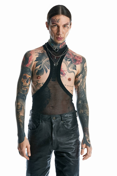 Молодой человек с обилием татуировок, украшающих его тело, уверенно позирует в студии на сером фоне. - Фото, изображение