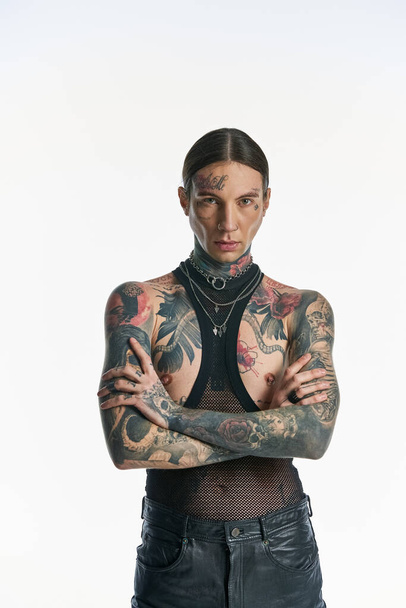 Ένας κομψός νεαρός με τατουάζ στέκεται με αυτοπεποίθηση, σταυρωμένα τα χέρια, σε ένα στούντιο πάνω σε γκρι φόντο.. - Φωτογραφία, εικόνα