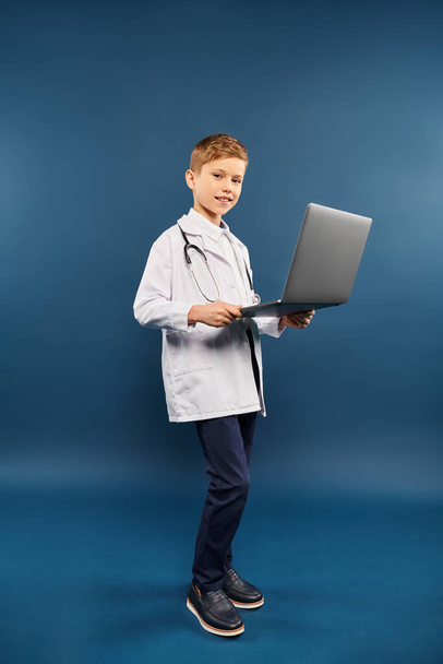 Un garçon préadolescent en blouse de laboratoire tenant un ordinateur portable sur fond bleu. - Photo, image