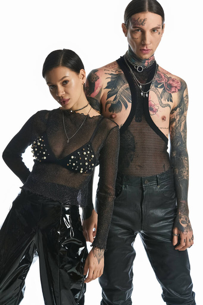 Egy fiatal, tetovált férfi és nő áll együtt egy stúdióban, szürke háttérrel.. - Fotó, kép