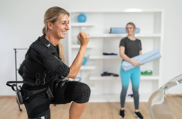 Nainen EMS-puvussa tekemässä kyykkyjä kouluttajan kanssa, jolla on joogamatto seuraavaan harjoitukseen EMS-studiossa - Valokuva, kuva