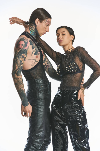 Ένας κομψός, με τατουάζ νεαρός άνδρας και γυναίκα στέκονται μαζί σε ένα στούντιο σε ένα γκρίζο φόντο, αποπνέοντας αυτοπεποίθηση και ενότητα. - Φωτογραφία, εικόνα