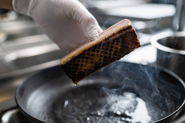 Kesztyűvel a kezében egy ropogós sertés has, amely sült egy forró olajos serpenyőben egy gáztűzhely egy professzionális konyhában egy étteremben. Luxus hotel főzés koncepció kép. - Fotó, kép
