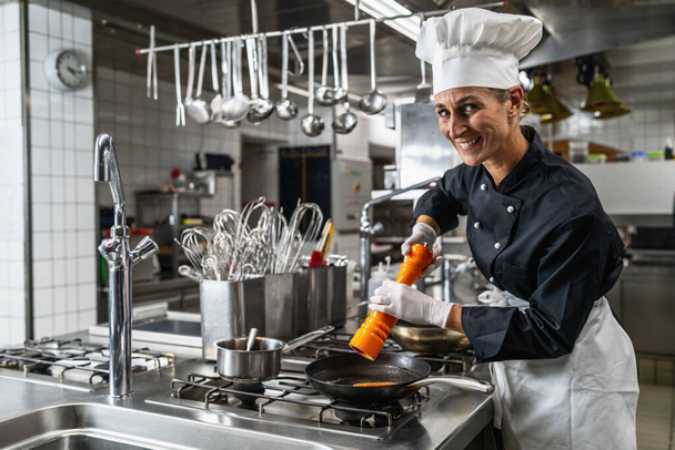 Chef feminino sorridente no hotel ou restaurante cozinha cozinhar e temperar a comida com um moinho de pimenta ou sal - Foto, Imagem