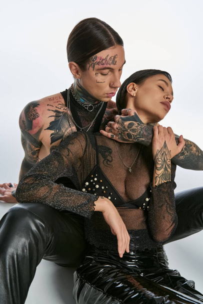 Ένας νέος, με τατουάζ άνδρας και γυναίκα κάθονται κοντά σε ένα στούντιο, αποπνέοντας στυλ και σύνδεση σε ένα γκρίζο φόντο. - Φωτογραφία, εικόνα