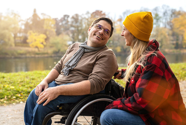 Άνδρας σε αναπηρική καρέκλα μιλάει με τη γυναίκα που τον φροντίζει σε ένα μονοπάτι, ενώ περπατούσε δίπλα στο ποτάμι το φθινόπωρο. Εικόνες έννοιας Joyful Connections - Φωτογραφία, εικόνα