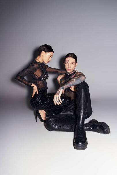 Dos mujeres jóvenes y elegantes con tatuajes sentadas juntas en un estudio sobre un fondo gris. - Foto, Imagen