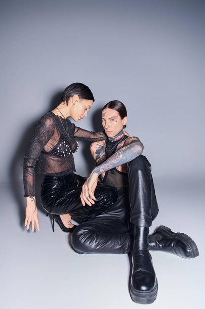 Молодой стильный мужчина и женщина с татуировками сидят бок о бок в студии на сером фоне. - Фото, изображение