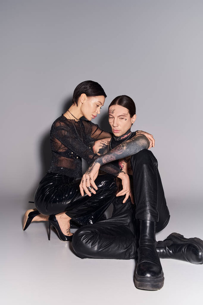 Un homme et une femme jeunes, élégants et tatoués assis ensemble sur un fond de studio gris. - Photo, image