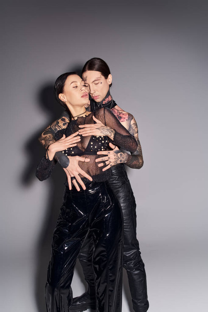 Ένα νεαρό κομψό ζευγάρι με τατουάζ αγκαλιάζονται θερμά σε ένα στούντιο με γκρι φόντο.. - Φωτογραφία, εικόνα
