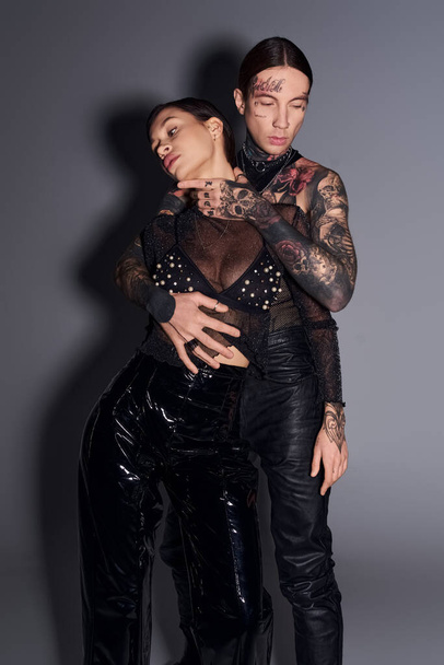 Egy fiatal, stílusos, tetovált pár áll együtt egy stúdióban, szürke háttérrel.. - Fotó, kép
