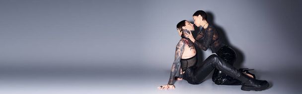 Una giovane donna elegante e tatuata siede aggraziatamente a terra vicino all'uomo in un ambiente da studio su uno sfondo grigio. - Foto, immagini