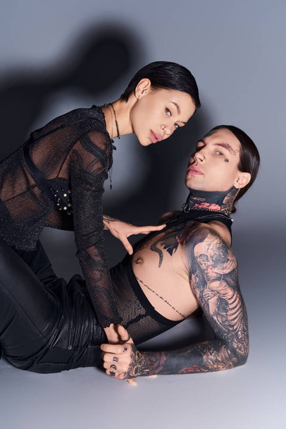 Ein stylischer junger Mann und eine junge Frau mit Tätowierungen posieren gemeinsam in einem Studio vor grauem Hintergrund. - Foto, Bild