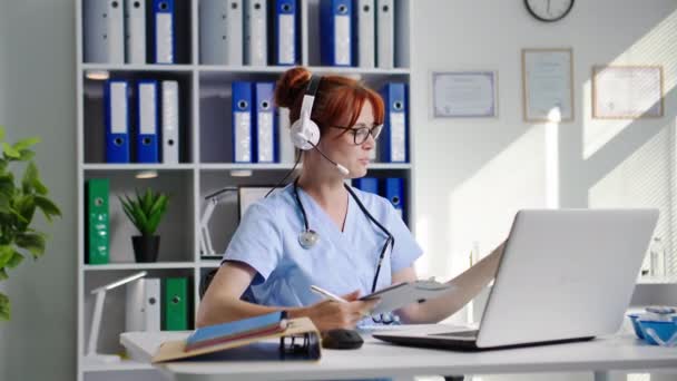 laboratorio en línea, asistente de laboratorio médico femenino con un auricular se comunica con el médico a través de videollamada en el ordenador portátil y muestra la reacción de los reactivos en el tubo de ensayo mientras está sentado en el consultorio médico - Imágenes, Vídeo