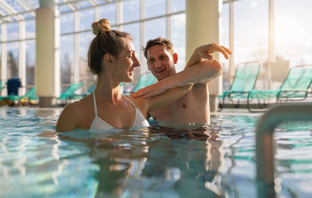 Treinador masculino auxiliando com exercício no ombro na piscina spa resort para hidroginástica na reabilitação - Foto, Imagem