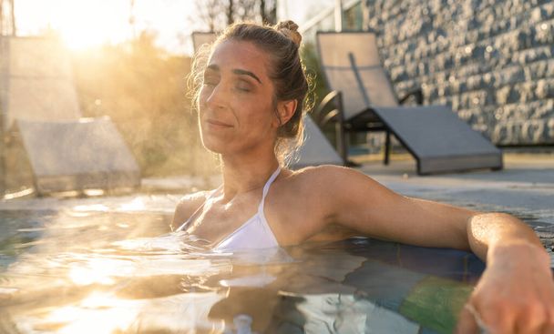 Женщина расслабляется в гидромассажной ванне с солнечной вспышкой, спокойное выражение лица в велнес-отеле - Фото, изображение