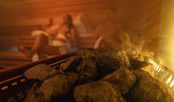 Vapor subindo de pedras de sauna com pessoas desfocadas no fundo, luz brilhante quente. relaxante em finlandês sauna spa hotel conceito imagem - Foto, Imagem
