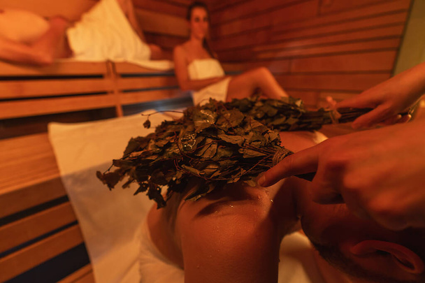 Close-up of a vihta (birch whisks) being used on a back of a man in a finish sauna, with others in the background - Photo, Image