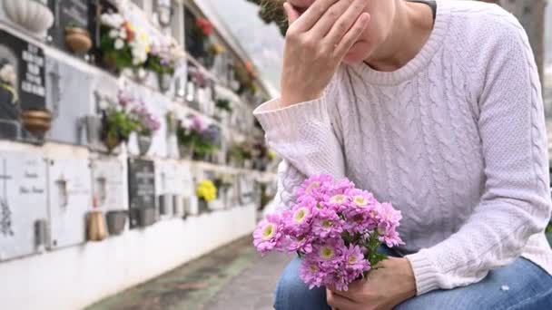  Oříznout truchlící samici v neformálním oblečení plačící a dotýkající se hlavy, zatímco sedí u hrobové zdi s čerstvými květy v ruce na hřbitově - Záběry, video