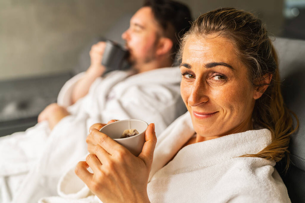 Mujer con una taza de té sonriendo a la cámara, hombre en bata de spa bebiendo té en el fondo en el centro de bienestar - Foto, Imagen