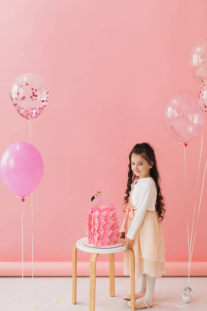 Rozkošná holčička v růžových šatech pózuje vedle plameňáka narozeninový dort a slaví své páté narozeniny na růžovém pozadí - Fotografie, Obrázek