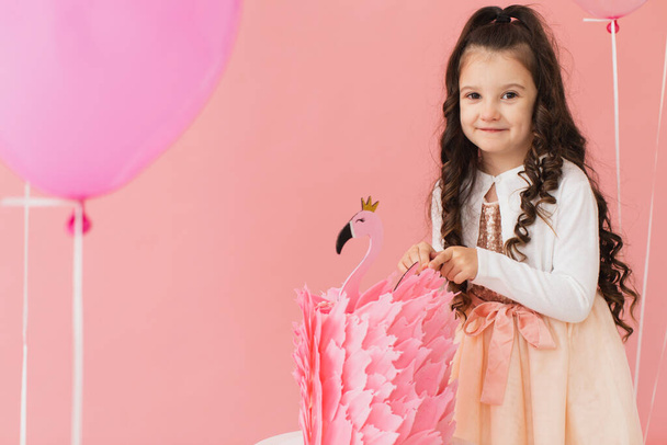Imádnivaló kislány rózsaszín ruhában, flamingó torta mellett pózol, és rózsaszín alapon ünnepli az ötödik születésnapját. - Fotó, kép