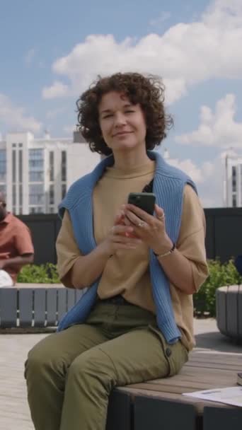 Függőleges teljes felvétel pozitív kaukázusi nő göndör haj ül padon a modern tetőtéri parkban mobiltelefon a kezében a napsütéses nyári napon, és nézi a kamera - Felvétel, videó