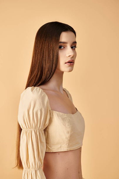 Μια νεαρή γυναίκα με μακριά καστανά μαλλιά, που ενσαρκώνει μια καλοκαιρινή διάθεση, ποζάρει με χάρη σε μια λευκή μπλούζα σε ένα σκηνικό στούντιο.. - Φωτογραφία, εικόνα