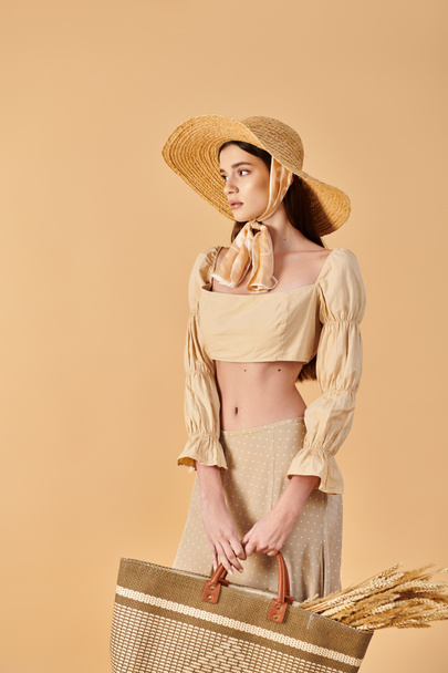 Młoda kobieta z długimi brunetkami pozuje spokojnie w letnim stroju, trzymając koszyk w stylowej czapce. - Zdjęcie, obraz
