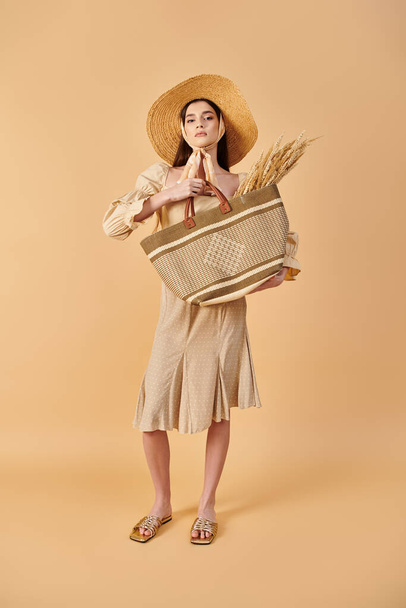 Μια νεαρή γυναίκα με μακριά καστανά μαλλιά ποζάρει κομψά σε ένα στούντιο, φορώντας καπέλο και κρατώντας μια τσάντα με καλοκαιρινή διάθεση. - Φωτογραφία, εικόνα