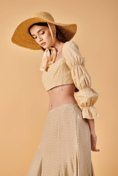 Модная молодая женщина с длинными брюнетками, ставящая позу в стильной шляпе и юбке, излучающая летнюю атмосферу в студии. - Фото, изображение