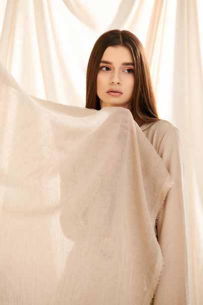 A young woman with long brunette hair strikes a pose in a summer outfit, exuding elegance and beauty in front of a white curtain. - Photo, Image