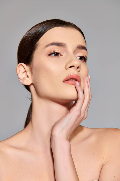 A young woman with brunette hair poses gracefully with her hand delicately resting on her face in a studio setting. - Photo, Image