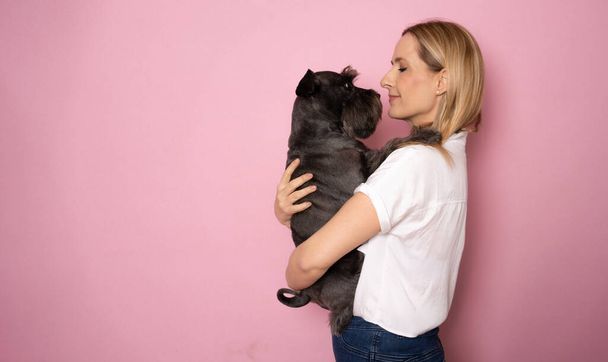 Cute young woman hugs her puppy schnauzer dog. Love between owner and dog. Isolated on pink background - Fotó, kép