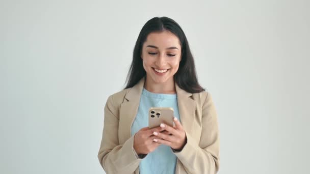 Pensive melko intialainen tai arabialainen nainen tyylikkäässä puvussa, matkapuhelimella viestimiseen ystävien kanssa sosiaalisessa mediassa, seisoo eristetyllä valkoisella taustalla, näyttää ajattelevalta, ajattelevalta, hymyilevältä - Materiaali, video