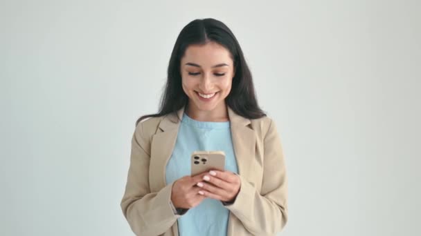 Використання гаджетів. Позитивна індійська або арабська жінка, одягнена в елегантний костюм, використовує смартфон для обміну повідомленнями з друзями, переглядає Інтернет, стоїть на ізольованому білому тлі, щасливо посміхається - Кадри, відео