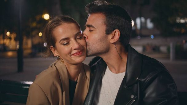 Gentle guy kissing girl cheek on date closeup. Smiling sweethearts in love resting urban park enjoy evening together. Relaxed stylish couple talk on street. Affectionate latino guy stroking girlfriend - Photo, Image