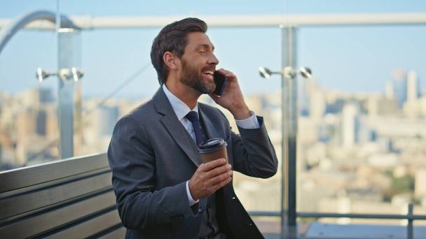 Empresario relajado hablando de teléfono móvil en la terraza. Feliz hombre barbudo hablando celular sosteniendo café en el paisaje urbano matutino. Alegre trabajador de oficina descansando balcón disfrutando de agradable conversación en el descanso - Foto, Imagen