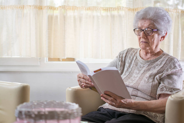 Oudere dame die een boek leest terwijl ze thuis in de woonkamer zit - Foto, afbeelding