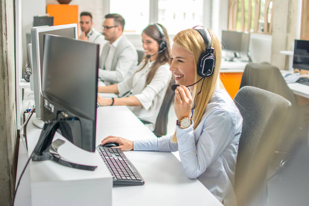 Call center operators.Young beautiful business woman with headset working on computer in office with colleagues in the background. - Photo, Image