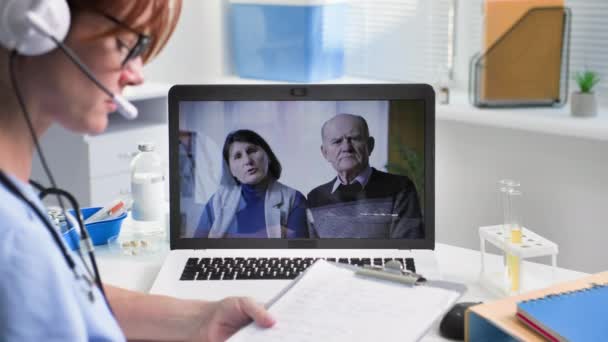 ontvangst van patiënten online, een jonge vrouwelijke arts communiceert met een oudere man en vrouw via videoconferentie op een laptop met behulp van een headset terwijl u in het medisch kantoor zit - Video