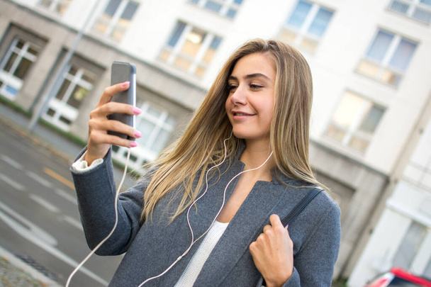 Lächelndes blondes Mädchen mit Smartphone, das auf der Straße Musik hört - Foto, Bild